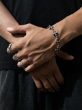 A close-up of a person's hands wearing a silver-toned chain bracelet with star and cross charms, along with several silver rings, against a dark black shirt background.